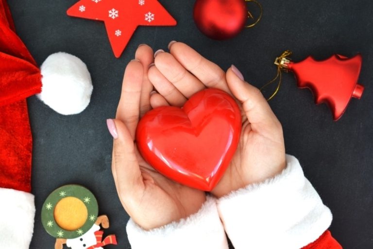 hands holding shiny red heart, wiht Christmas decorations around.